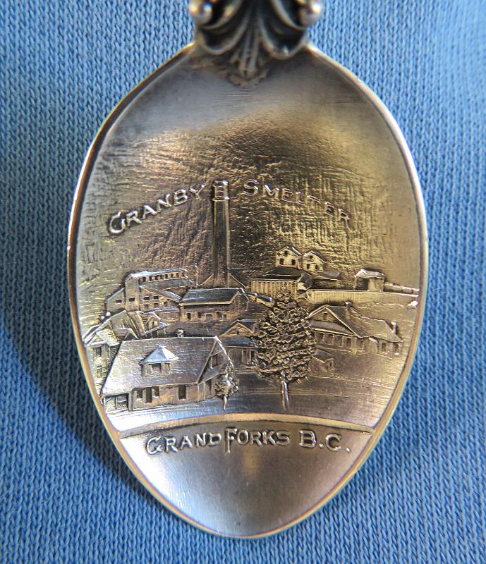Souvenir Mining Spoon Granby Smelter Bowl.JPG - SOUVENIR MINING SPOON GRANBY SMELTER GRAND FORKS BC - Sterling silver souvenir demitasse spoon, features handle with enamel BC crest at top, bowl embossed with detailed scene of the Granby Smelter and marked GRANBY SMELTER GRAND FORKS, B.C., length 4 in., marked on reverse Sterling with registered design RD 1901, weight13.8 gms. [The Granby Smelter was constructed in 1900 by the Granby Consolidated Mining and Smelting Company at Grand Forks, British Columbia.  Grand Forks is a city in the Boundary Country of the West Kootenay region of British Columbia. It is located at the confluence of the Granby River and Kettle River, a tributary of the Columbia. The city is just north of the US-Canada border. Grand Forks was established in the late 19th century when copper mining dominated Boundary and Kootenay regions of BC. The city was laid out in 1895 and Grand Forks was officially established as a city on 15 April 1897. The Phoenix Mine was an open pit and underground copper mining operation near the town of Grand Forks.  It was a lower grade copper deposit that was discovered in 1891.  In 1896, J.F.C. Miner, a rubber footwear manufacturer from Granby, Quebec, and mining promoters J. P. Graves and A. L. Little formed the Miner-Graves Syndicate and purchased the Phoenix Mine. In 1899, they incorporated The Granby Consolidated Mining and Smelting Company and, in 1901, consolidated as Granby Consolidated Mining, Smelting and Power Company. In spite of the low grade of the ore from the Phoenix deposit, it had the advantage of being self-fluxing (only requiring coke to be added for smelting), which resulted in a cheaper processing cost. In 1896 development of the deposit began and in 1900 the Granby Smelter Company completed the 700-ton-per-day smelter in Grand Forks which would at the time be the largest non-ferrous smelter in the British Empire and the second largest in the world.  Ore was transported to the smelter via the Canadian Pacific Railway, which had built a line to the mining operation.  By 1910 the smelter had undergone several expansions whereby it employed 300 men and processed 3000 tons of ore per day.  With the dramatic drop in world copper prices in 1919, the smelter and Phoenix mine closed.  The slag piles on the Granby River just outside town are all that remains today of this large copper smelting operation.]